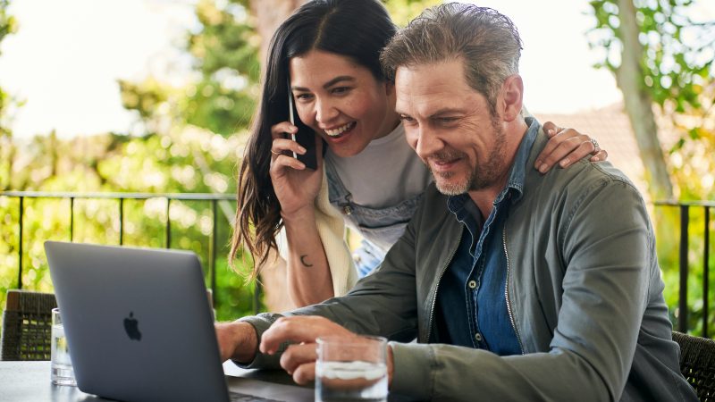 couple qui se trouve devant leur ordinateur en extérieur, l'homme écrit sur le pc et la femme est souriante au téléphone
